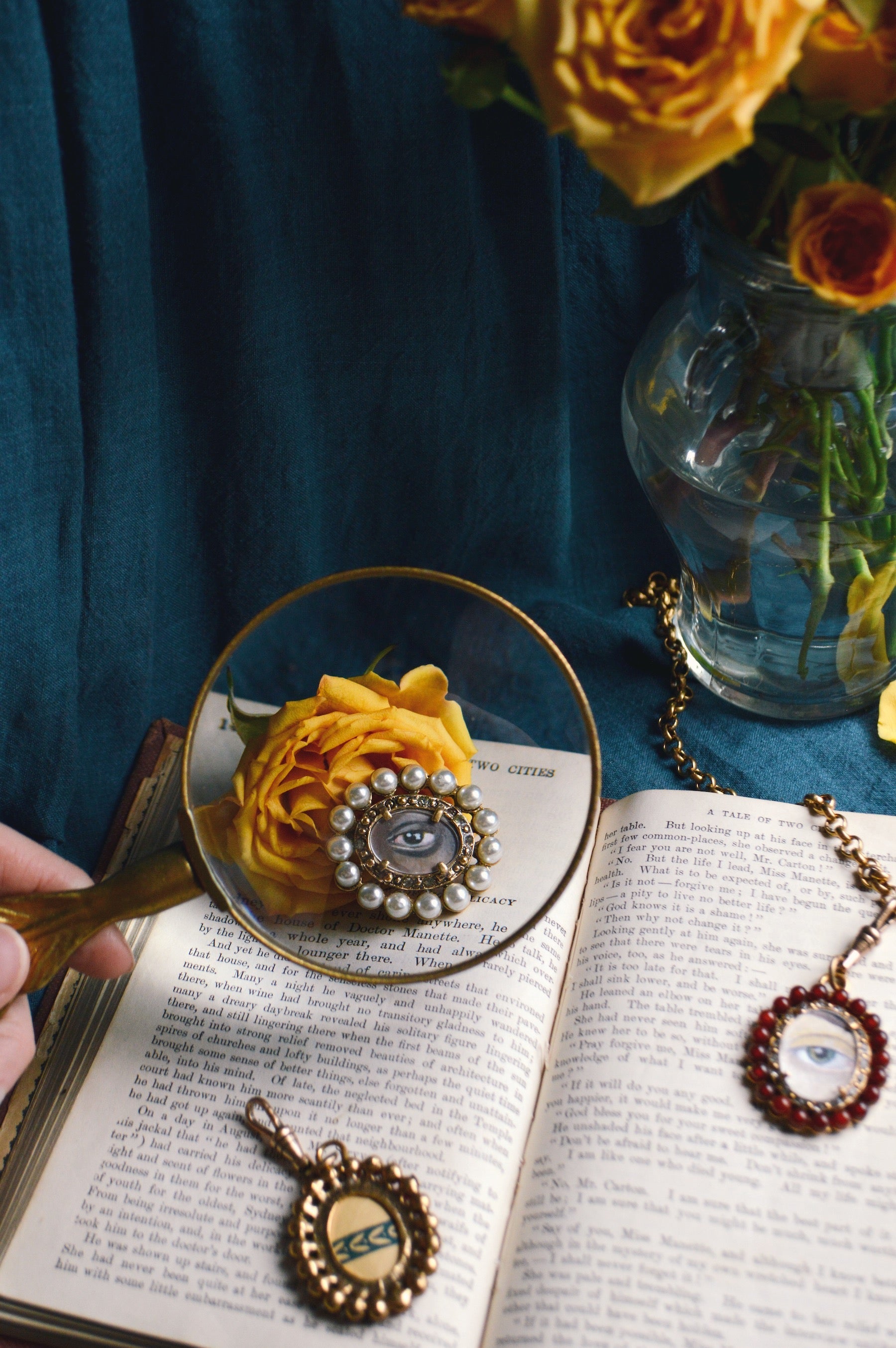 Against a dramatic, deep blue background, golden roses rest in a glass vase and on top of an antique book open to display French text. A white person's hand holds a magnifying glass over the rose and book, and at its center is the enlarged image of a hori