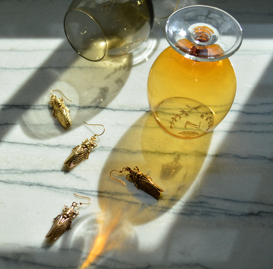 A scattering of four, golden cicadas with detailed wings and heads rests over dark-veined white marble, two amber-colored beer glasses perched above with sunlight streaming through.